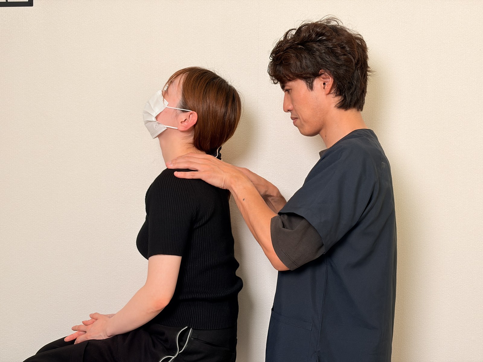 座った姿勢で首を前に倒してもらい、肩に手を添えて動く範囲を確認する院長